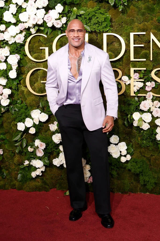 The Rock on red carpet wearing pastel blazer, silk shirt, and dark trousers in front of floral backdrop with &quot;Golden Globes&quot; sign