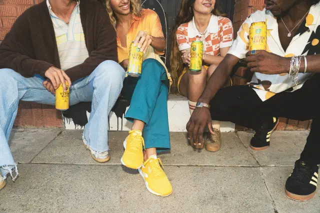 Four people sitting on a sidewalk, each holding a can, wearing casual summer outfits and sneakers