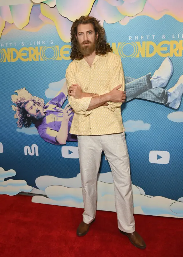 Rhett on red carpet wearing a patterned shirt and light pants, standing in front of a whimsical poster backdrop