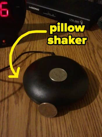 Black electronic pillow shaker on a wooden surface, shown next to coins for size comparison