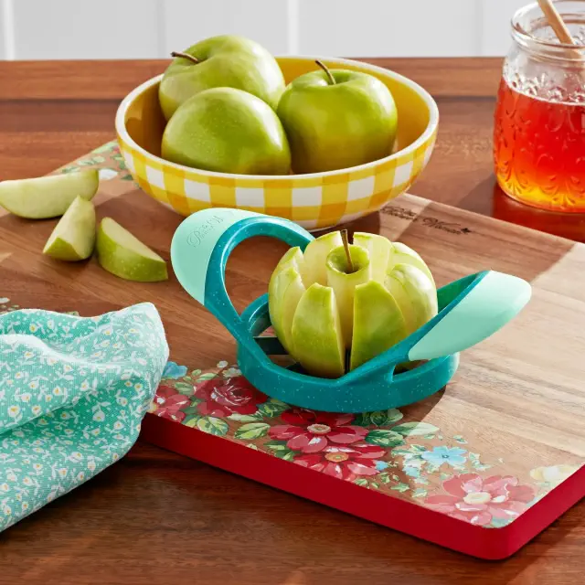 Apple slicer cutting a green apple on a decorative cutting board, with whole apples in a bowl and a jar of honey nearby