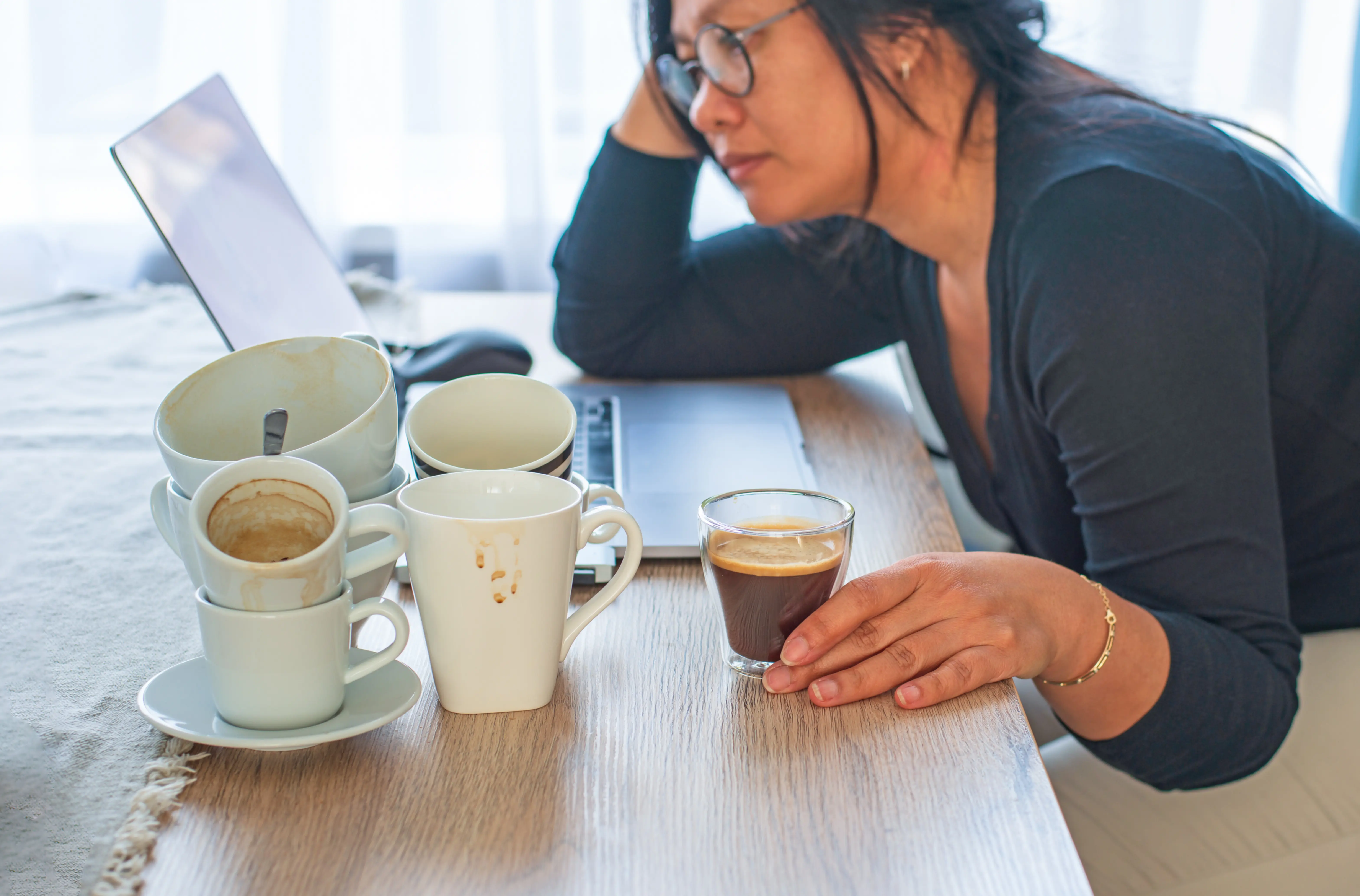 A woman sits at a table resting her head on her hand, reading her laptop. She is surrounded by empty coffee cups. 
