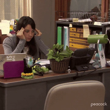 Woman at desk exasperatedly holds head in hands, surrounded by office supplies and a plant, while phone rings