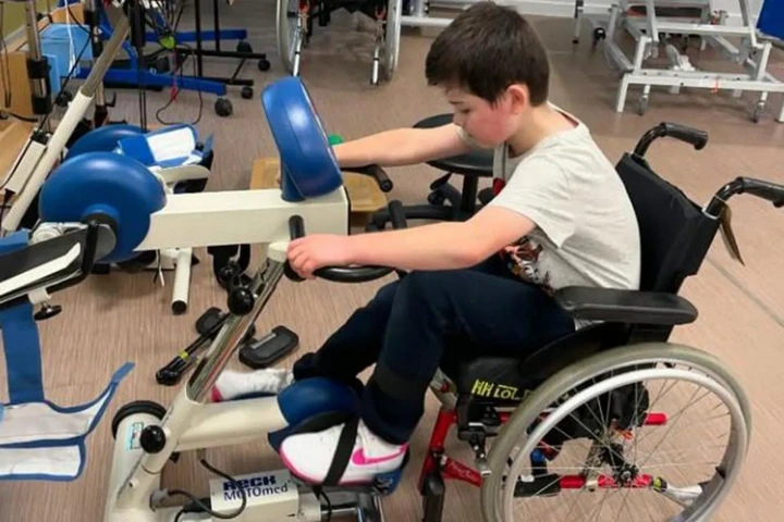 Family pamphlet/PA Real Life Harry is seen in a wheelchair using a physical therapy exercise machine as part of a rehabilitation room with other equipment in the background. 