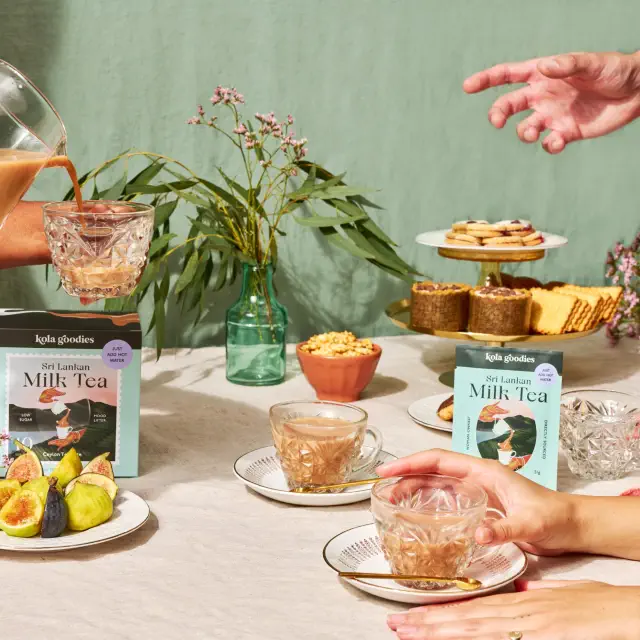 Milk tea is being poured into glass cups on a table with snacks and pink flowers in the background. Packages of milk tea are displayed