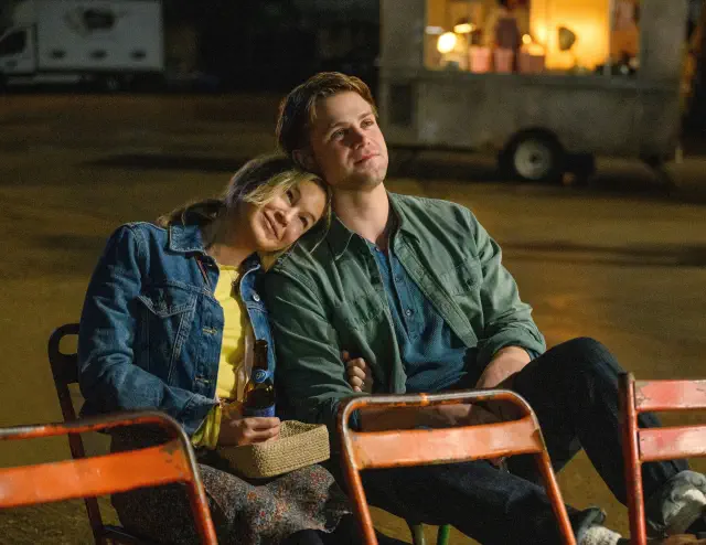 Two people sit closely on chairs, one resting their head on the other&#x27;s shoulder, appearing to enjoy a quiet, intimate moment together outdoors
