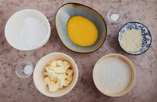 Ingredients in bowls: powdered sugar, melted butter, flour, egg yolks, almond flour, salt, and baking powder, arranged on a countertop