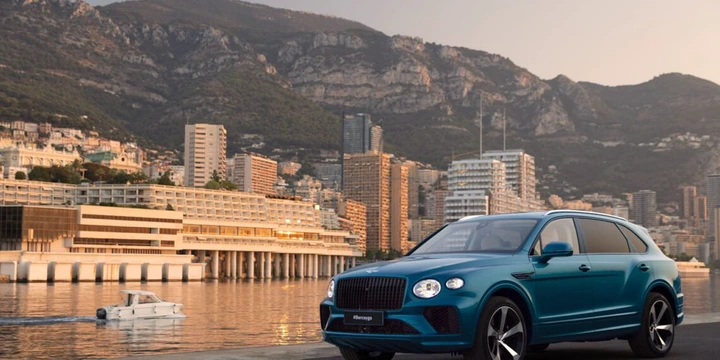 A blue Bentley Bentayga from the EWB Riviera Collection is parked by a waterfront with a city skyline and mountains in the background. A white boat glides through the water, creating a picturesque scene reminiscent of Brussels' finest views.