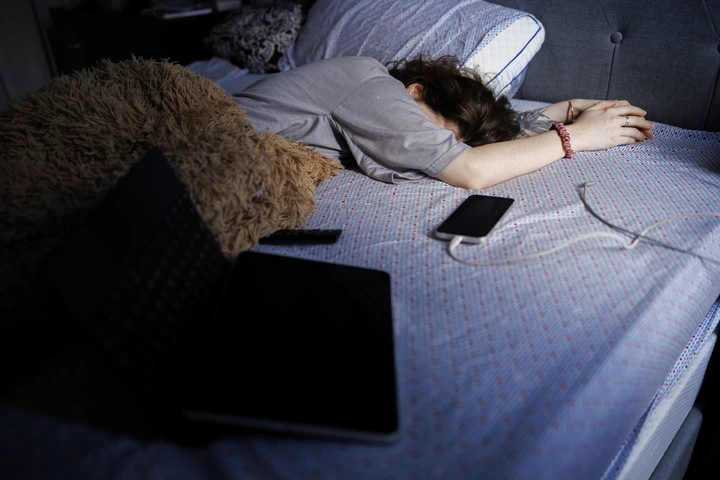 Person lying face down on a bed with a phone and an open laptop nearby, suggesting rest or exhaustion