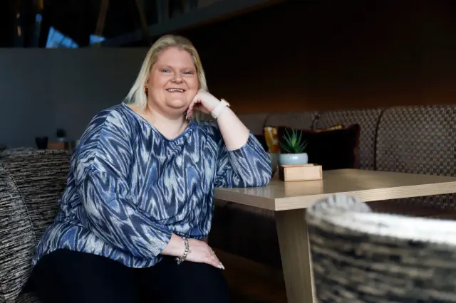 Person sitting in a modern lounge area, wearing a patterned top and black pants, smiling at the camera