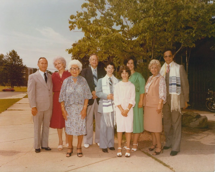 A group of people pose outdoors, dressed in formal attire. The central person wears a tallit, suggesting a special occasion