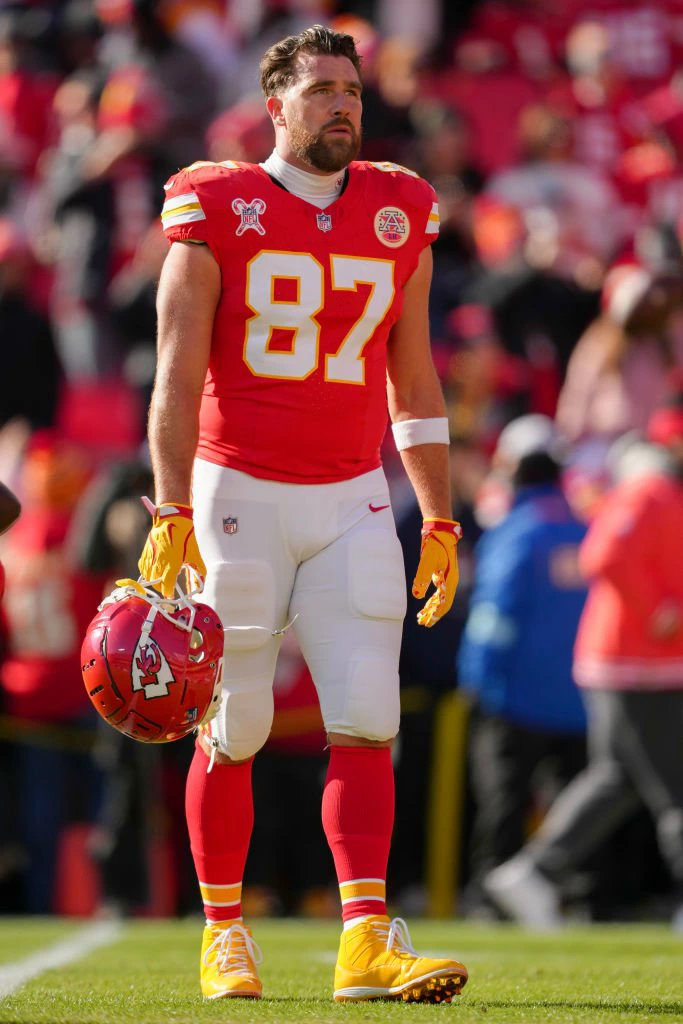 Travis in a football uniform, holding his helmet, stands on the field