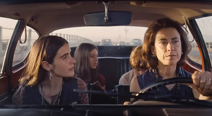 Four people in a car: a woman driving, a young woman in the passenger seat, and two children in the back. The mood is serious and contemplative