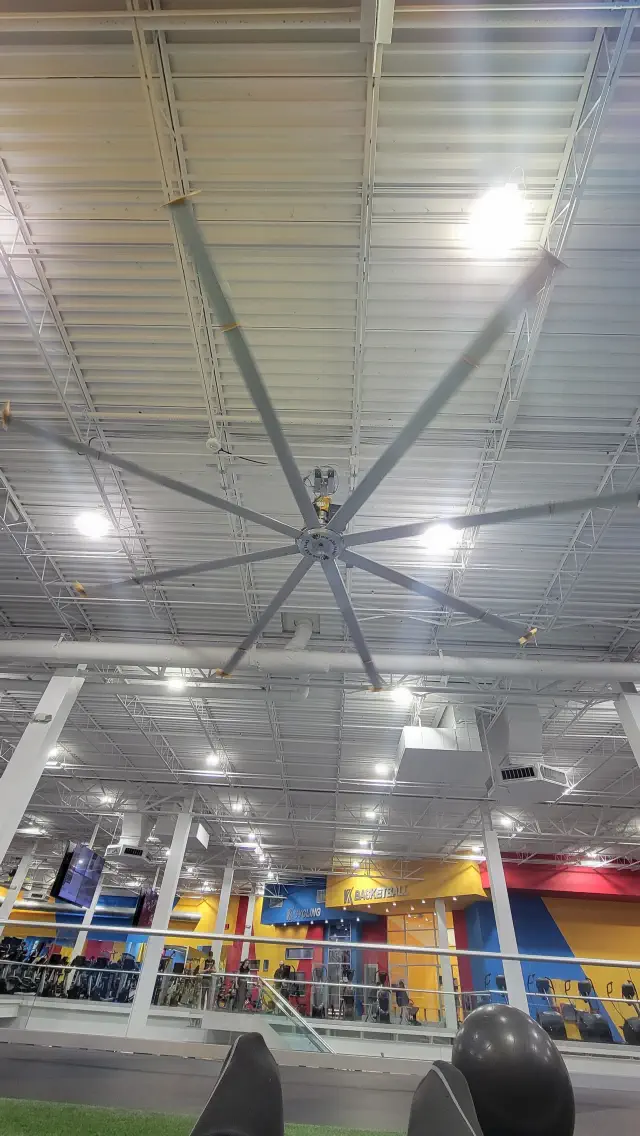 View of a large ceiling fan in a gym with a high ceiling and exercise equipment in the background