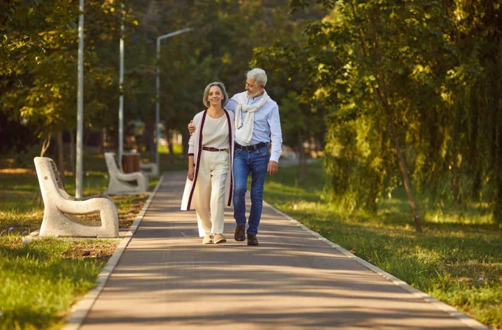 An elderly couple, husband and wife, stroll hand in hand through a city park, relishing their vacation together. They appear lively and joyful, embracing their time off as they meander and converse in the fresh air.