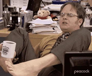 Man in office setting with feet on desk, holding a coffee mug, surrounded by cluttered papers and office supplies. Assistant TV show scene