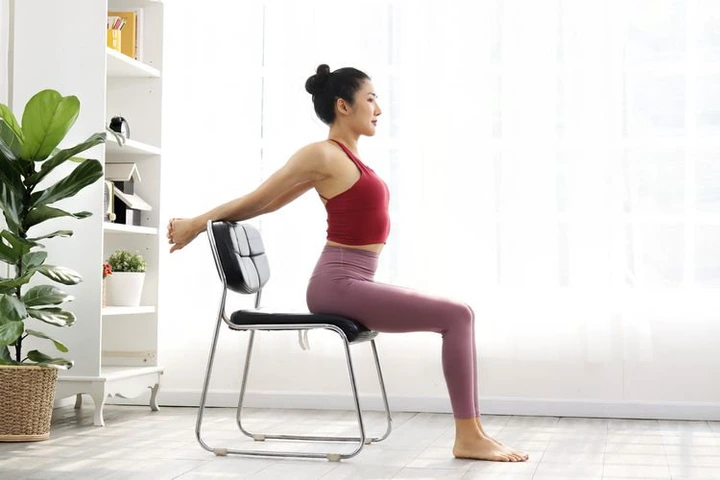 A young woman in athletic wear exercising at her desk using a chair. Engaging in stretches to combat the office blues.
