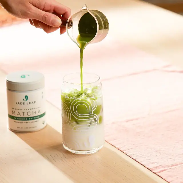 A hand pouring matcha into a glass of milk beside a container of Jade Leaf matcha, on a table
