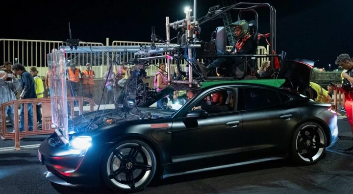 A camera rig is securely mounted on a sleek Porsche Taycan, with a crew member seated atop during an intense nighttime film shoot for Den Of Thieves 2, surrounded by bustling crew members in the background.
