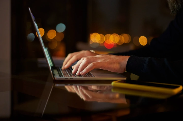 Person typing on a laptop at night, with blurred city lights in the background, suggesting a work-related activity