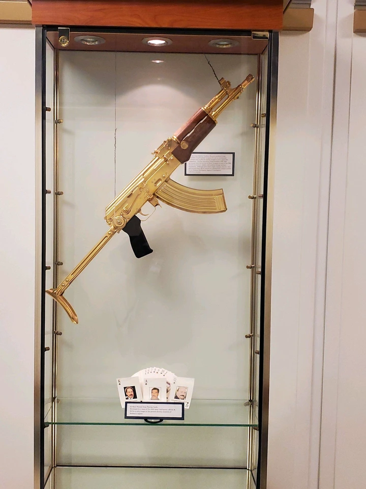A gold-plated AK-47 rifle displayed in a glass case with photos and cards beneath it