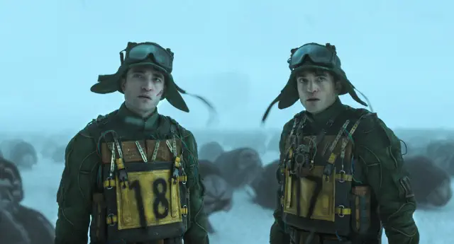 Two individuals in military-style gear with large goggles and helmets stand in a snowy landscape. They have numbered vests: 18 and 17