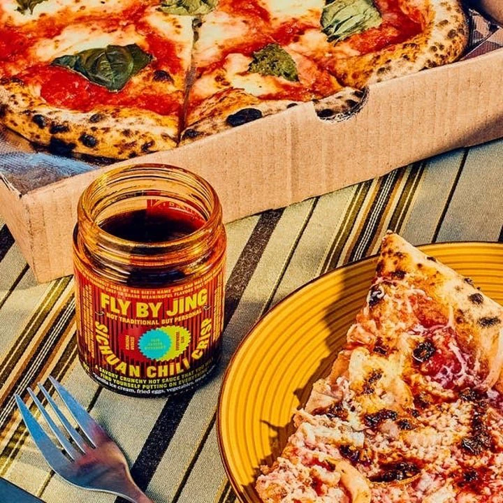 A jar of Fly By Jing Sichuan Chili Crisp is placed next to a slice and a box of pizza on a table with a fork nearby