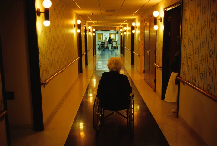 An elderly person in a wheelchair is in a nursing home hallway, moving toward a well-lit area with staff members in the distance