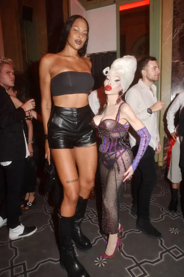 Liz and Amanda at a celebrity event: Liz wearing a strapless top and leather shorts, Amanda in a rhinestone bodysuit with a dramatic hairstyle. In heels, Amanda comes up to Liz&#x27;s chest