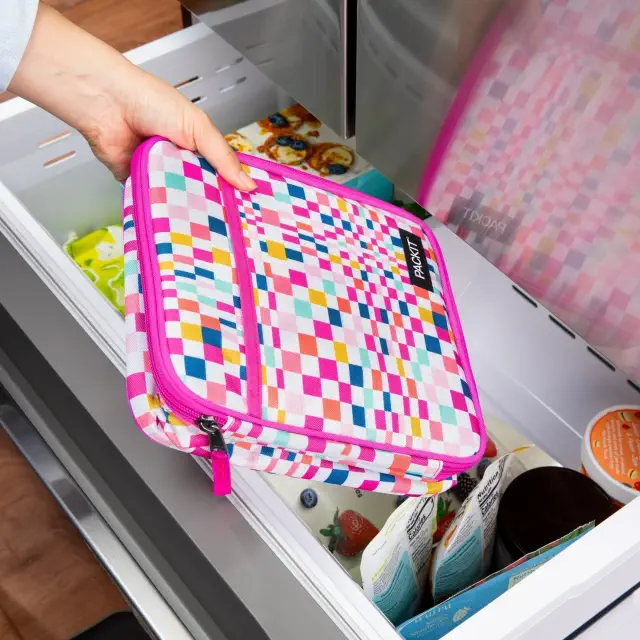 A person places a colorful, checkered lunch bag into a freezer drawer, filled with groceries. The bag is made by PackIt