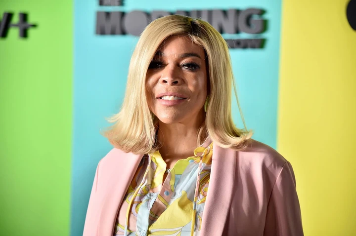 Wendy Williams smiling against 'The Morning Show' blue, green, and yellow background. She has shoulder-length blonde hair and she's wearing a pink coat over a bouse.