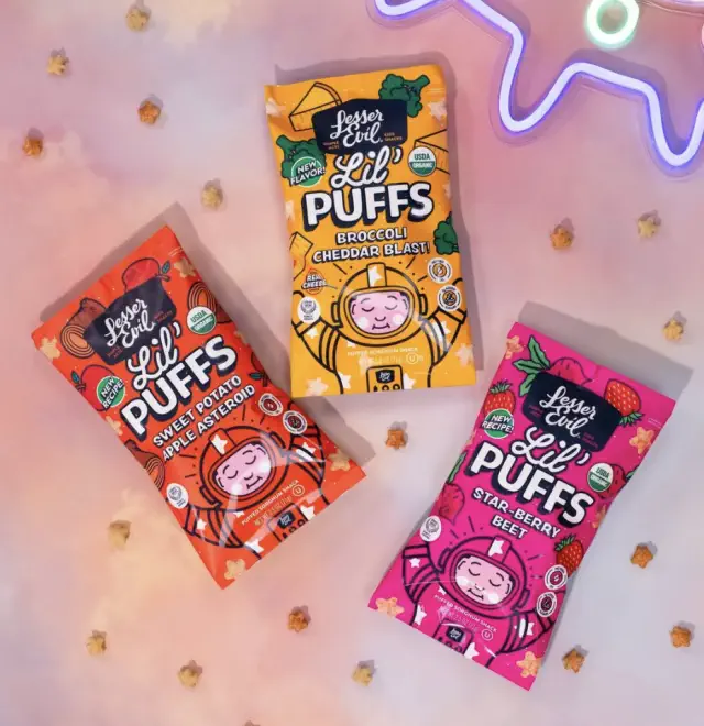 Three bags of Lesser Evil Lil Puffs snacks are displayed, featuring flavors like Sweet Potato Apple, Broccoli Cheddar, and Star Berry. Playful packaging
