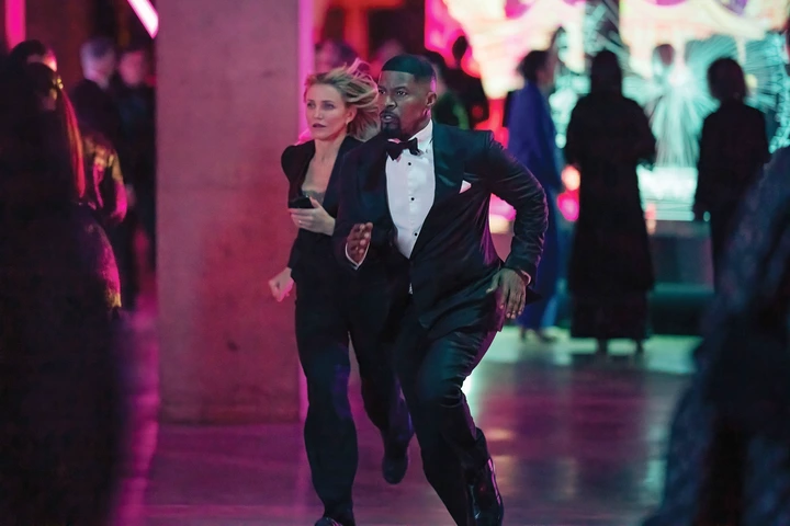 A woman in a tailored suit and a man in a tuxedo run through a lively, elegant event space