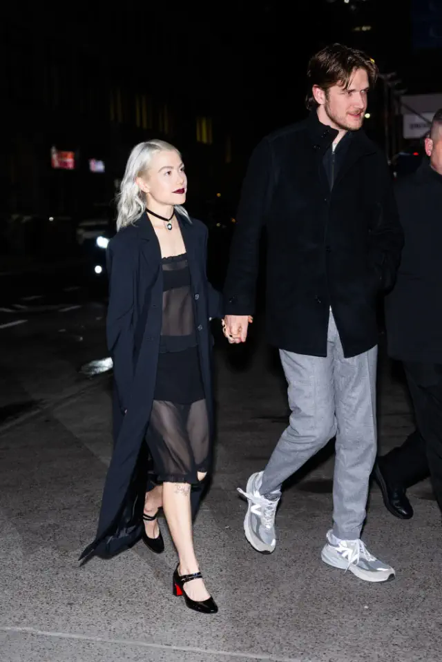 Phoebe and Bo walking together. She wears a sheer dress with a long coat and heels. He wears a dark jacket and light pants. In heels, she does not reach his shoulder