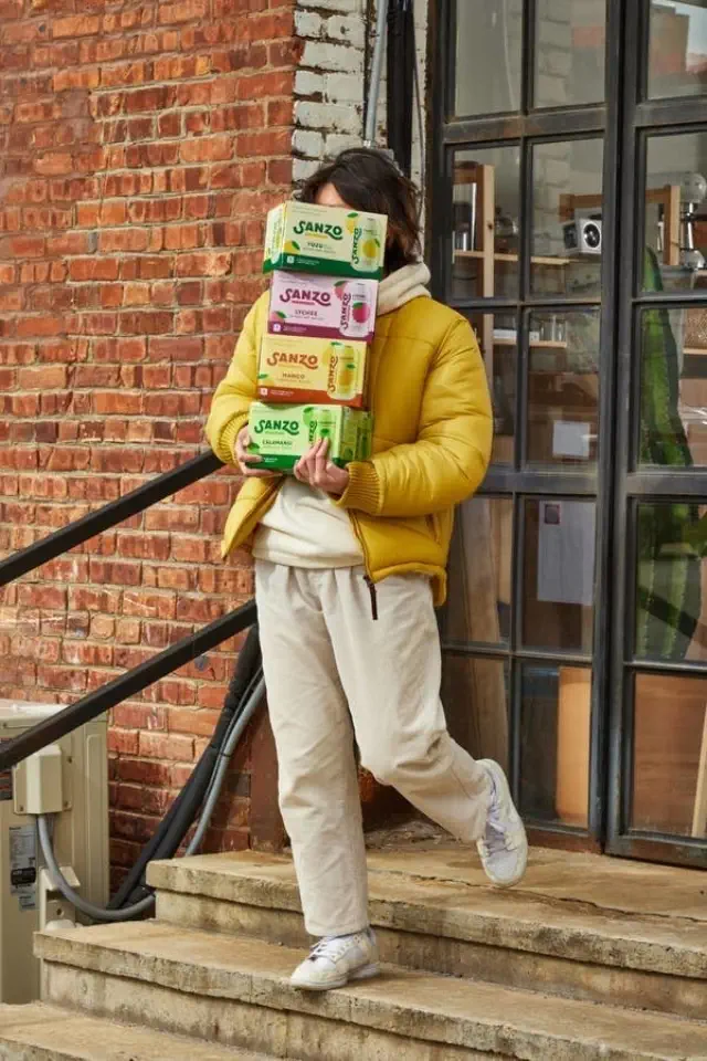 Person holding four boxes of Sanzo drinks while walking down concrete stairs outside a brick building. Wearing a yellow puffer jacket and beige pants