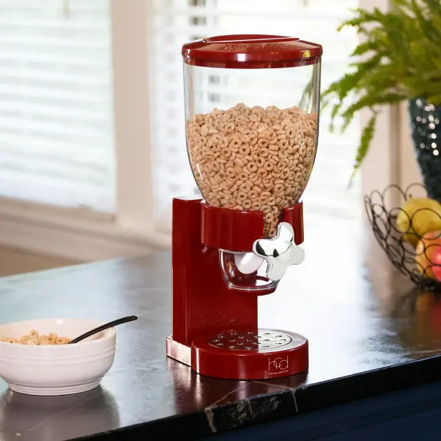 Cereal dispenser on a kitchen counter next to a bowl and fruit basket
