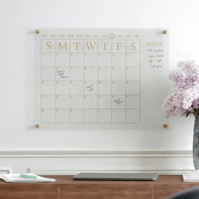 A wall-mounted glass calendar with handwritten notes and reminders for the month, placed above a desk with a laptop and a vase of flowers