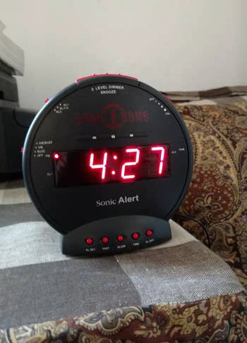 Digital alarm clock on a patterned bedspread, showing the time 4:27