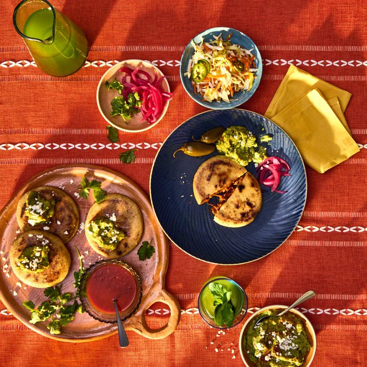 A vibrant table setting with pupusas, salsa, pickled onions, and slaw on decorative plates and a striped tablecloth