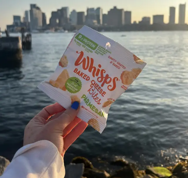 Person holding a bag of Whisps Baked Cheese Bites with a city skyline and water in the background