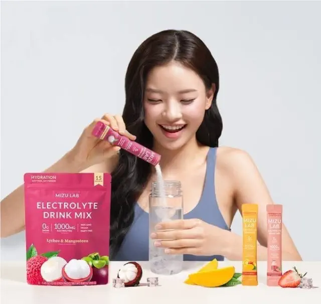 A smiling woman in a tank top is pouring Mizu Laboratory's electrolyte drink mix into a bottle. The drink mix flavors pictured are lychee &amp; mangosteen and orange