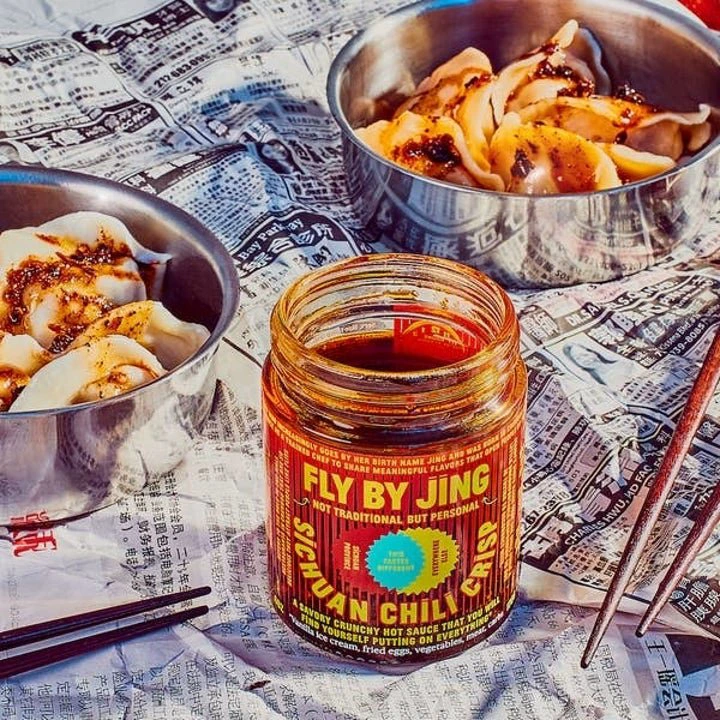 Fly By Jing's Sichuan Chili Crisp jar on a table with two bowls of dumplings covered in sauce, placed on a newspaper backdrop with chopsticks nearby