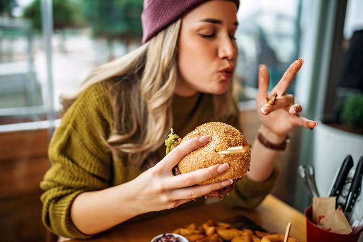 Person enjoying a large burger with sesame seed bun, seated at a table with fries, wearing a beanie and sweater, savoring the meal