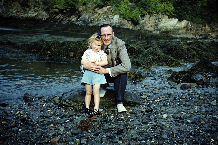A man and a young child stand on a rocky shore by water, smiling at the camera. The child is in a floral top and blue shorts, and the man is in casual attire