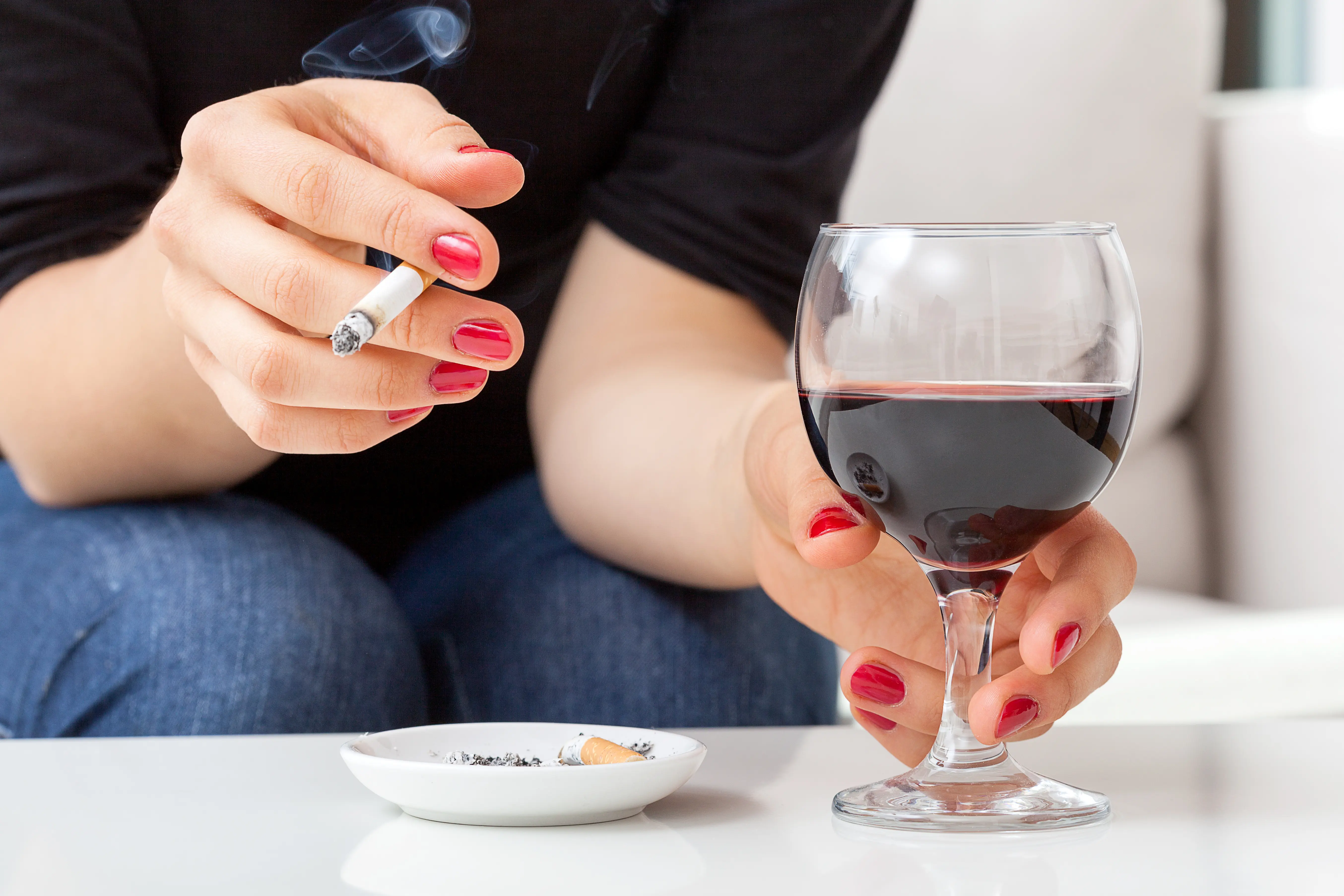 A woman enjoys a class of red wine while smoking a cigarette. 