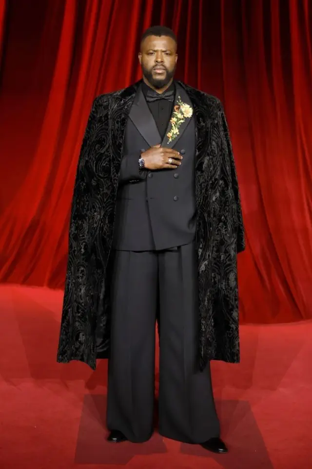 Winston on a red carpet in a tuxedo with a matching patterned cape, wearing a floral brooch, looking directly at the camera