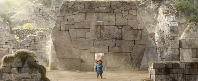 A small bear wearing a duffle coat and hat stands in front of ancient stone ruins, looking up at a large carved face on the wall