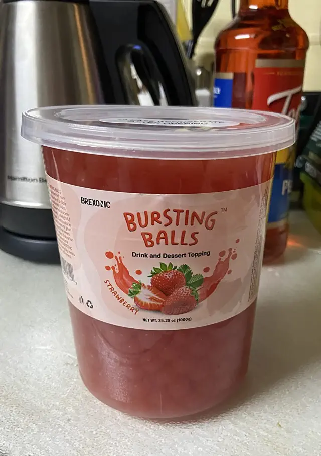 A container of Bursting Balls strawberry-flavored drink and dessert topping sits on a kitchen counter next to various kitchen items