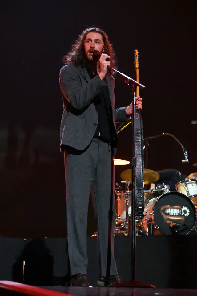 Hozier in a dark suit speaking into a microphone on stage, holding a guitar. Drums are visible in the background