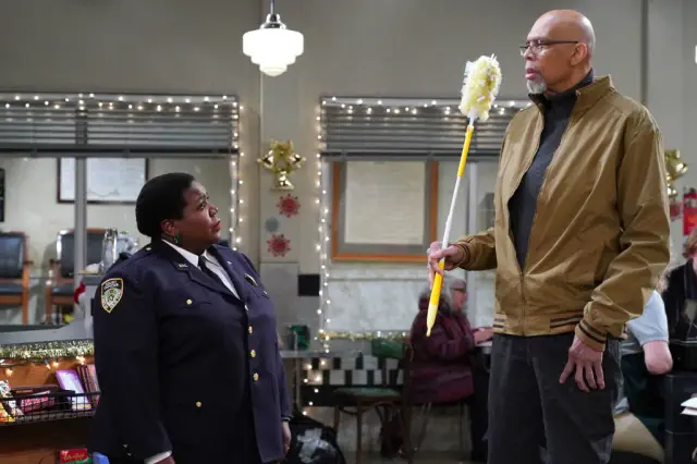 Lacretta and Kareem in a comedic scene: her in a police uniform looking up at him holding a mop in a casual jacket. She doesn&#x27;t reach his chest